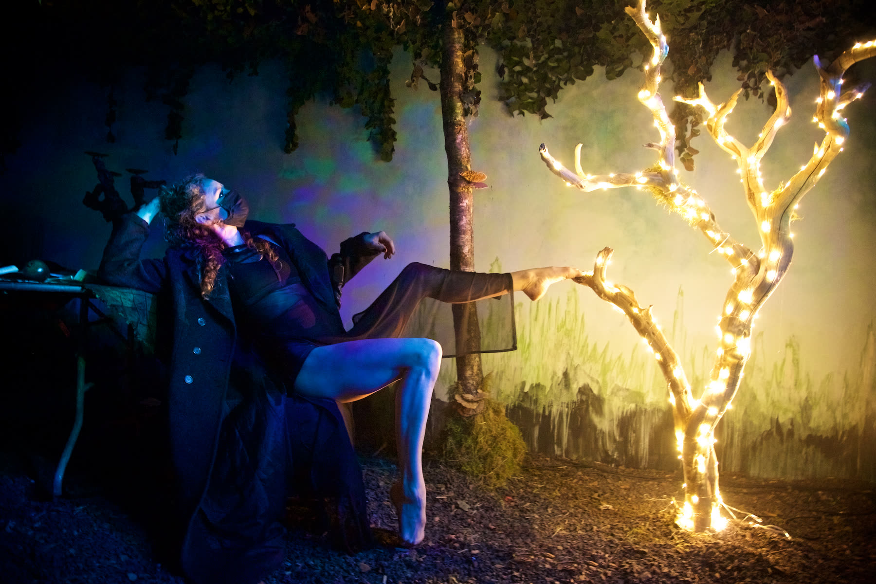 A costumed performer in character beside an illuminated tree on a painted set