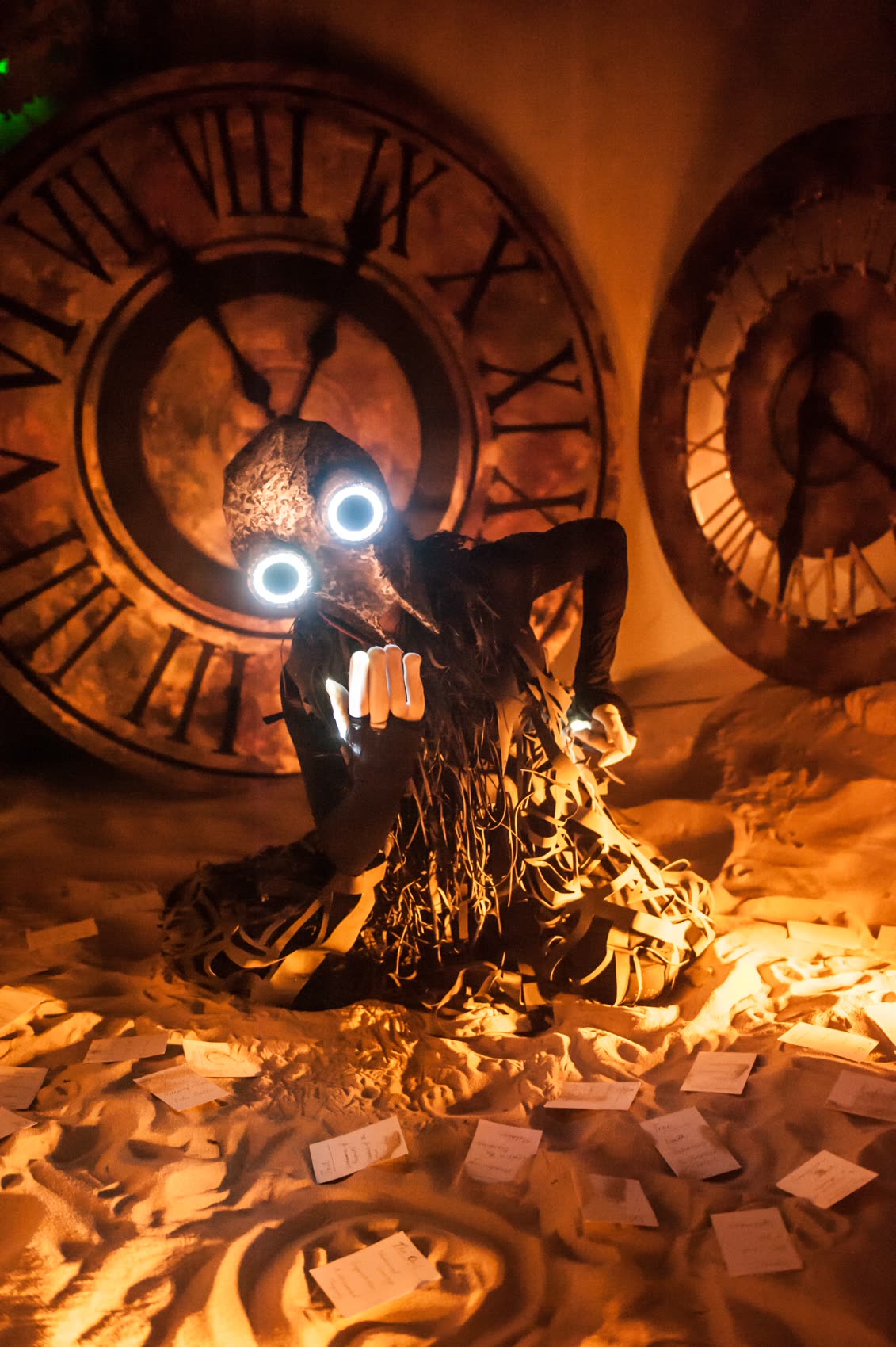 A masked performer with glowing eyes seated in sand in front of oversized clock faces